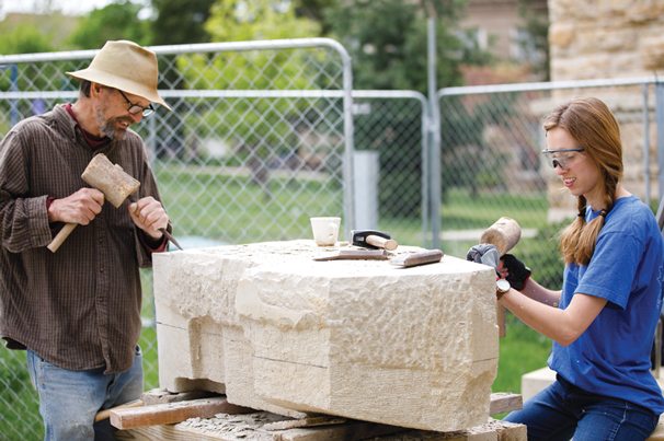 CARVING STATION: Sculptor Karl Ramberg uses hand tools to shape a 1,600-pound slab of Cottonwood limestone into a blocky animal-like shape that will eventually make its way to Laura Ramberg for the final carving. In 2019, Karl Ramberg worked with students, including Mattie Karr (pictured here), during the outdoor carving process in front of Dyche Hall.

Photo by University of Kansas/Andy White